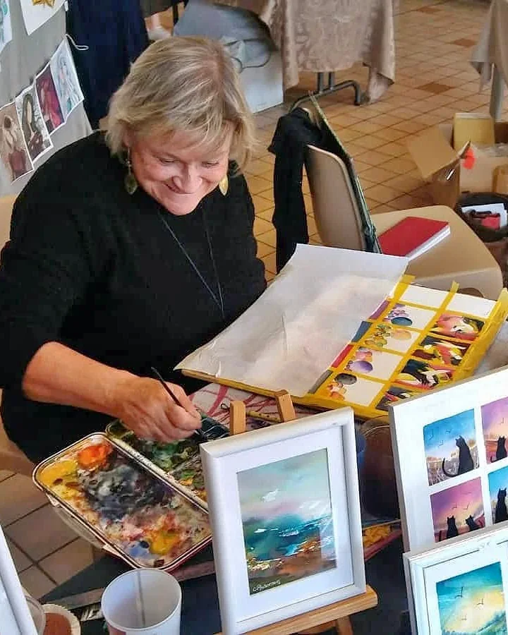 Chantal Anderson peint une aquarelle sur son stand, entourée de ses tableaux